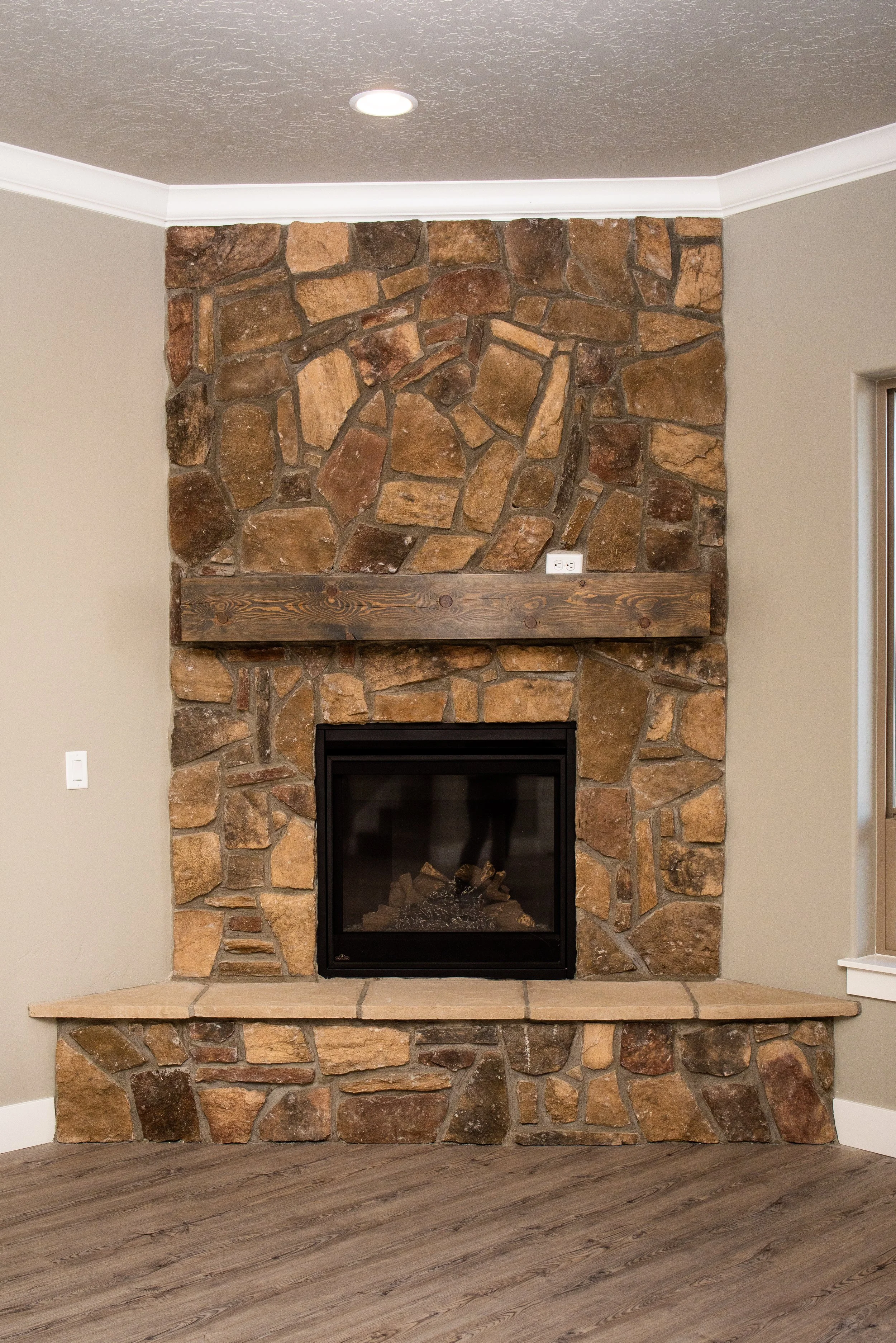 A stone fireplace with a dark firebox in a living room, on a beige tiled hearth, with beige walls and a wood-look flooring.