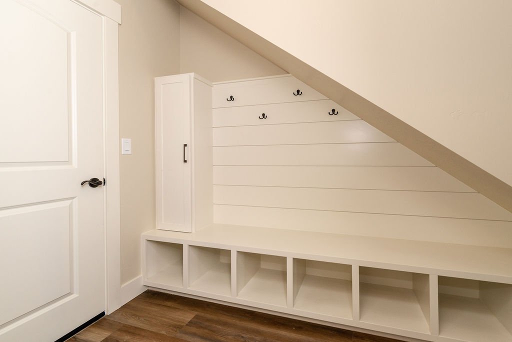 Wall nook with built-in white storage bench and hooks, under a sloped ceiling, next to a white door with a black handle.