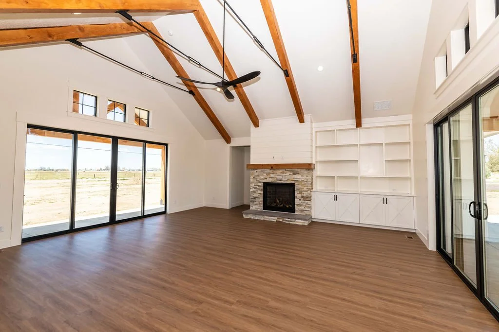 Empty living room with wood floors, white walls, a stone fireplace, built-in white shelves, large sliding glass doors, and exposed wooden ceiling beams.
