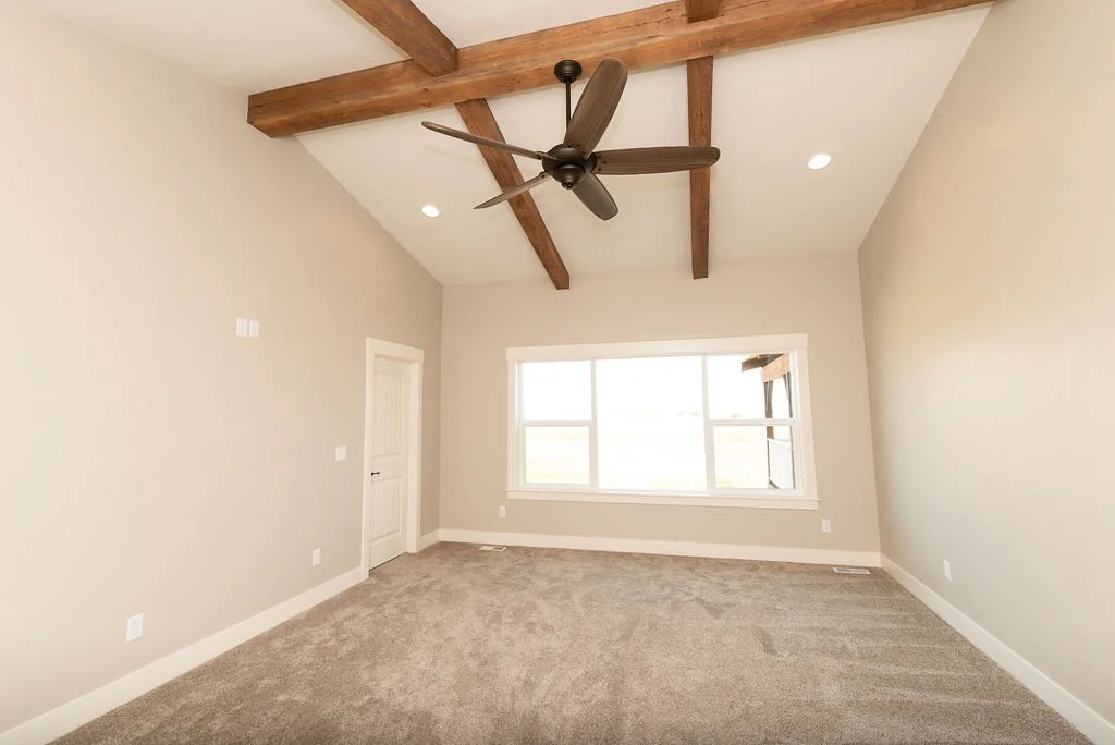 Empty living room with beige walls, large window, carpeted floor, and wooden ceiling beams with a ceiling fan.