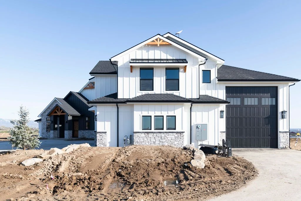 Newly constructed two-story house with white exterior, black roof, and attached garage, surrounded by dirt and construction materials.