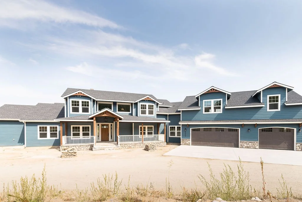 A large blue modern house with multiple gable roofs, white-framed windows, a covered front porch, and a three-car garage. The house is situated in a yard with dirt and sparse vegetation.