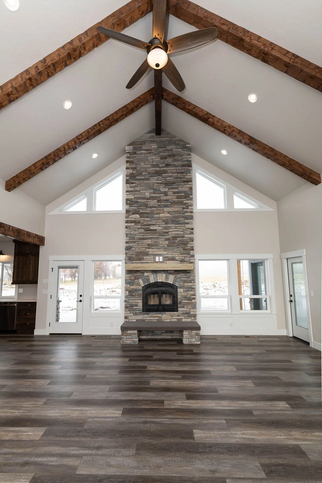 Interior of a spacious living room featuring a high vaulted ceiling with exposed wooden beams, a stone fireplace with a black wood stove insert, and large triangular windows. The room has a modern rustic style with hardwood flooring and neutral-color