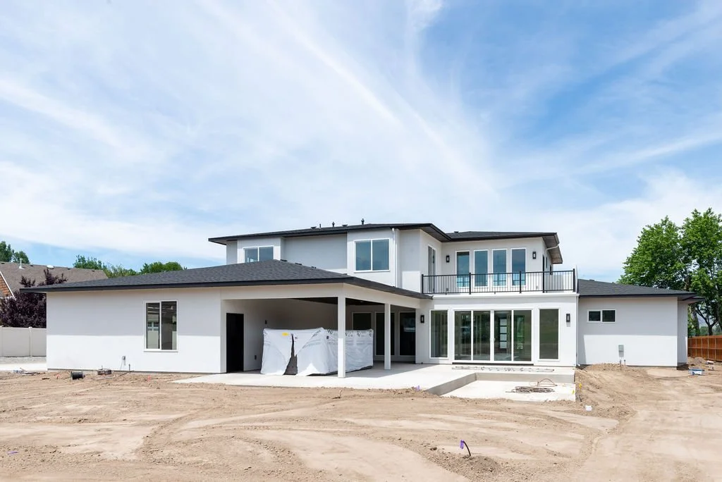 Under construction modern two-story house with white exterior, large windows, and a balcony, in a dirt yard with a blue sky.