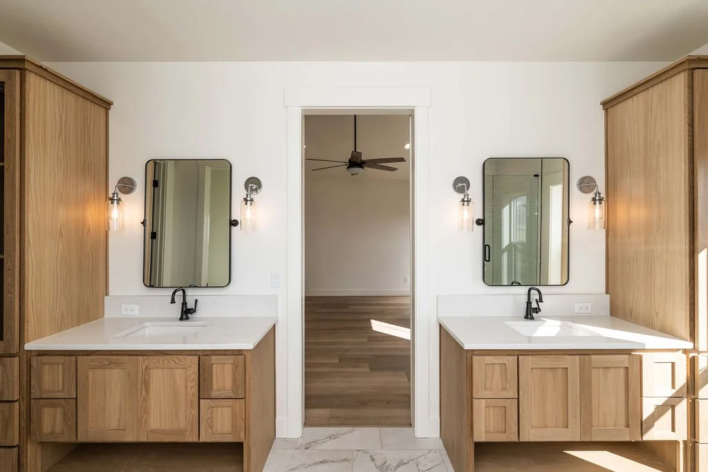 Dual bathroom vanities with white countertops, black faucets, large wall mirrors, and wall-mounted light fixtures, set against wooden cabinetry and a doorway leading to a room with hardwood floors and a ceiling fan.