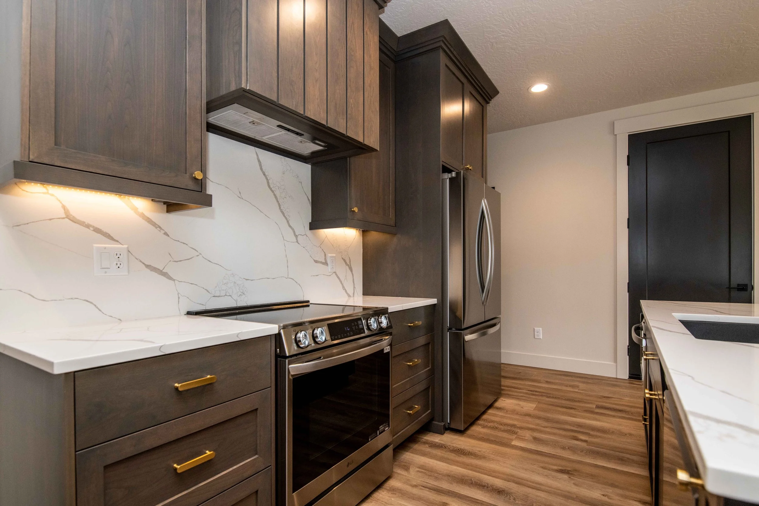 A modern kitchen with dark wooden cabinets, white marble countertops, stainless steel appliances including a stove and refrigerator, and hardwood flooring.