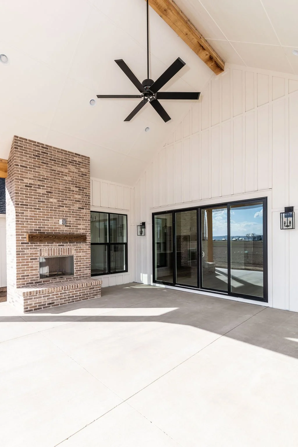 Outdoor patio with a brick fireplace, sliding glass doors, and black window frames under a vaulted ceiling with a ceiling fan.