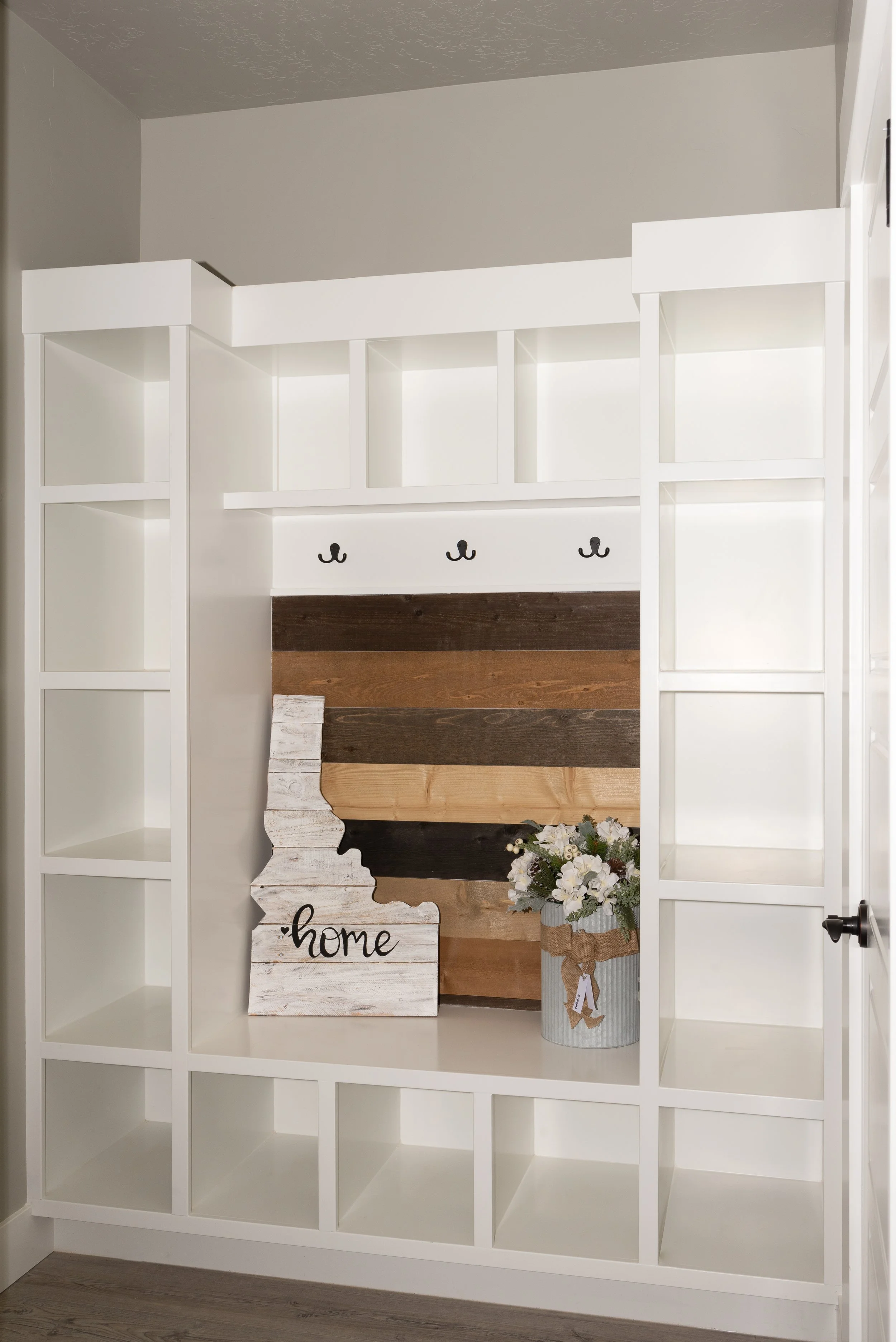 White shelving unit with cubbies, decorated with a wooden country-style 'home' sign, a bouquet of white flowers in a gray container, and a wooden plank wall with a plush animal and embroidery hoop.