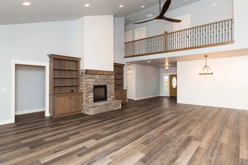 Empty living room with wood flooring, brick fireplace, built-in wooden shelves, white walls, high ceiling with ceiling fan, and an upstairs balcony with wooden railing.