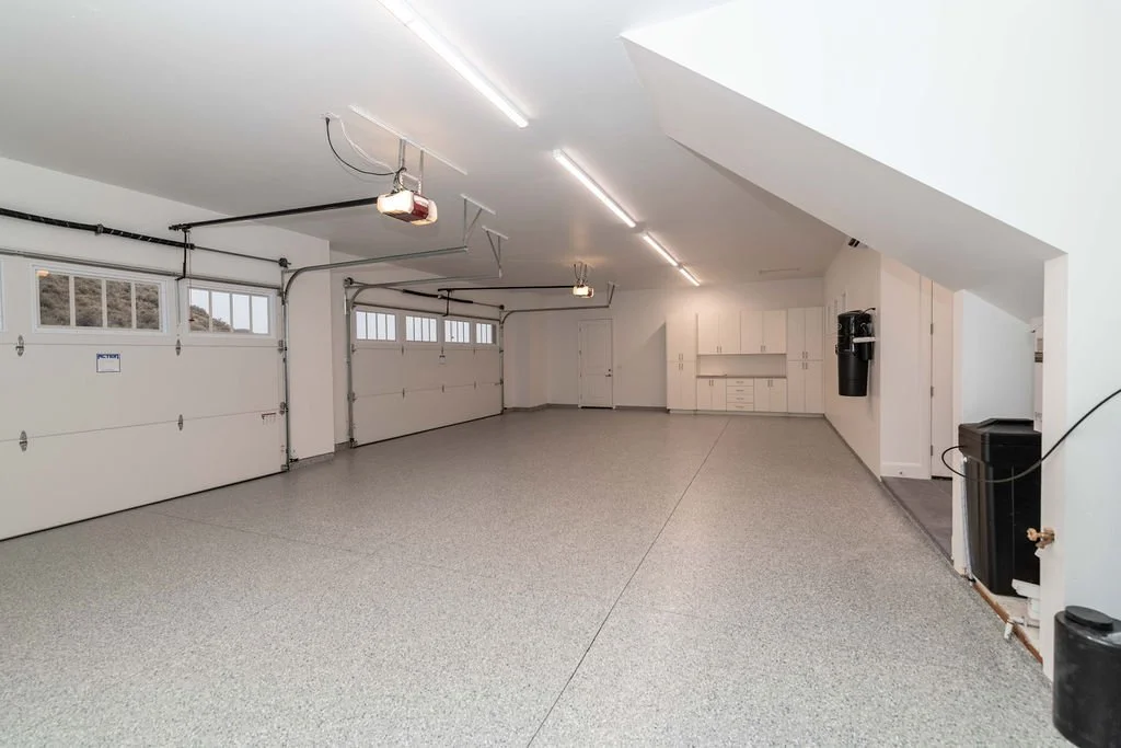 Empty, large two-car garage with white walls, speckled epoxy floor, ceiling lights, overhead garage door openers, and storage cabinets along the back wall.