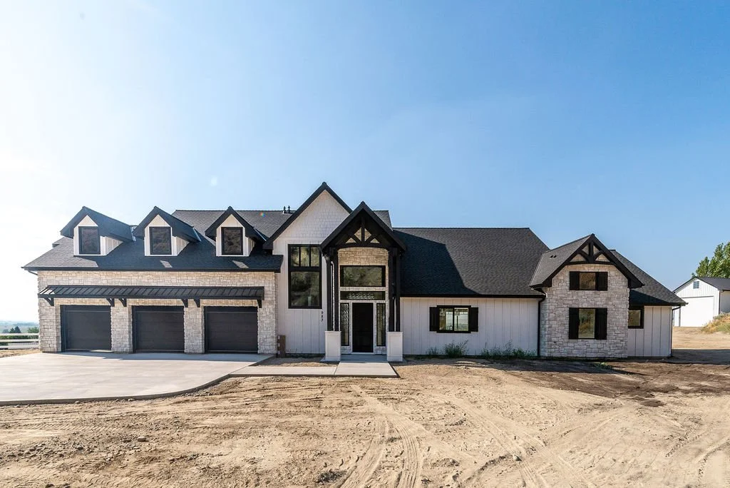 Newly built modern house with multiple garage doors, black window frames, and a combination of brick and white siding, set on a dirt lot with a clear blue sky.