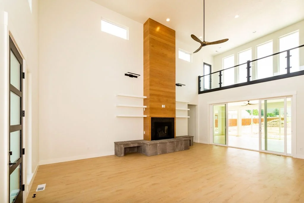 Bright living room with high ceiling, large windows, sliding glass doors, modern ceiling fan, and a fireplace with a wooden chimney and concrete hearth.