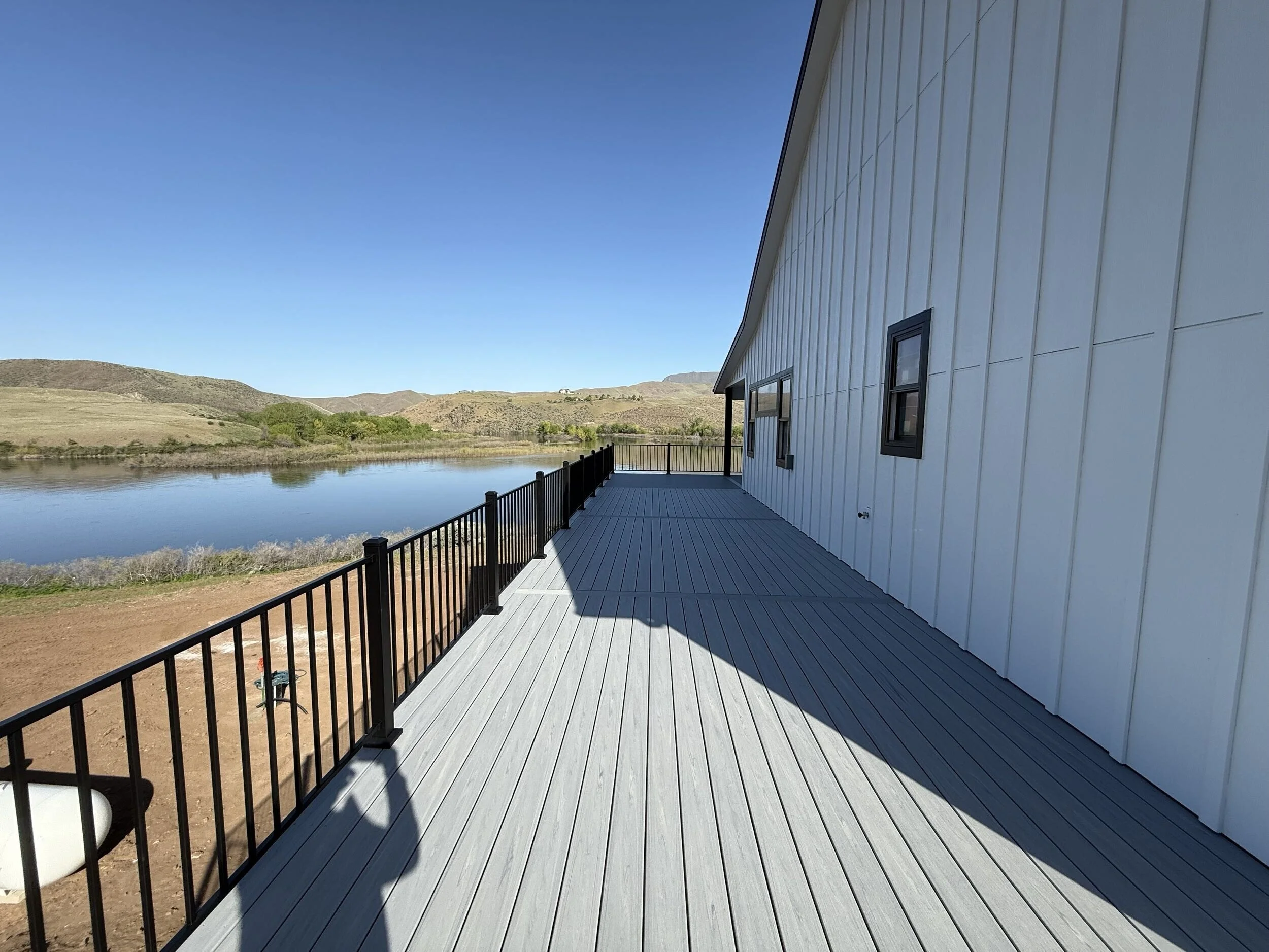 A wooden deck attached to a white building with black-framed windows, overlooking a body of water and rolling hills under a clear blue sky.