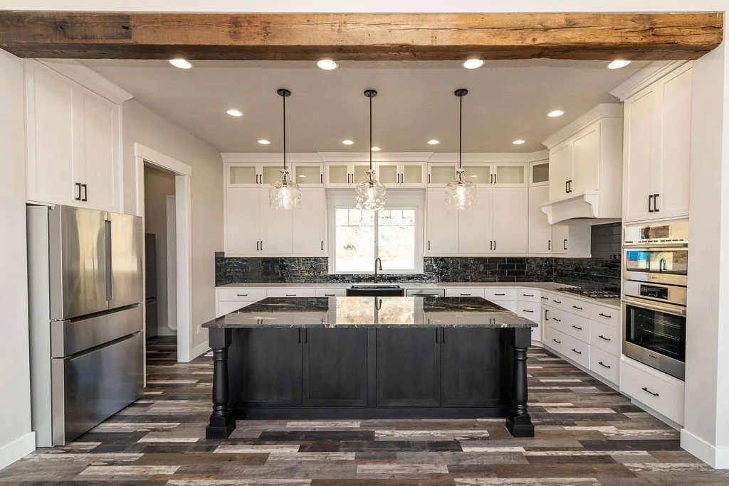 Modern kitchen with white cabinets, black countertops, a large island with black cabinetry, stainless steel refrigerator, and a window above the sink. Wooden beams on the ceiling and pendant lights above the island.