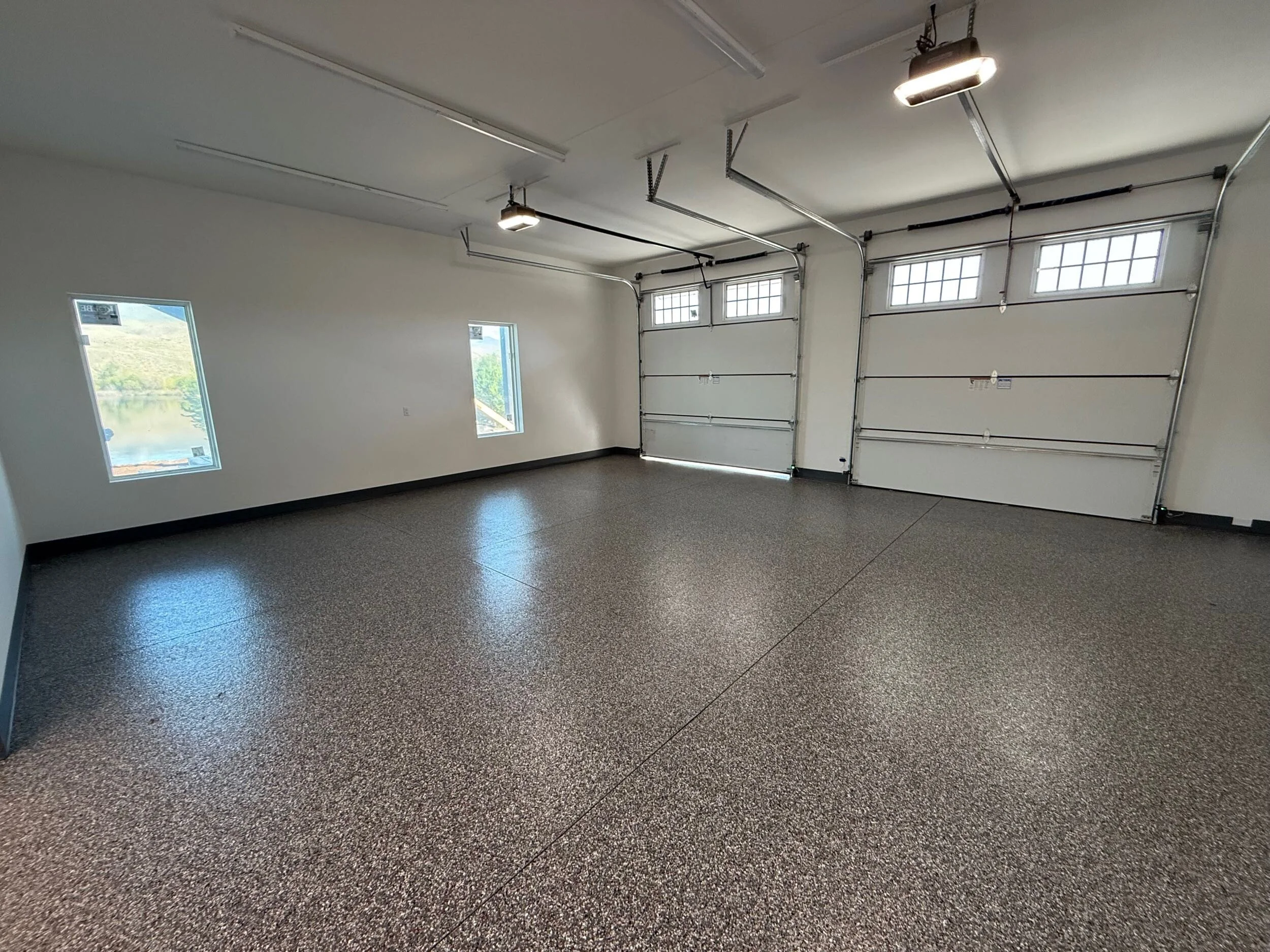 An empty garage with white walls, three windows, and a gray speckled epoxy floor. The garage has a white overhead door with windows and a garage door opener mounted on the ceiling.