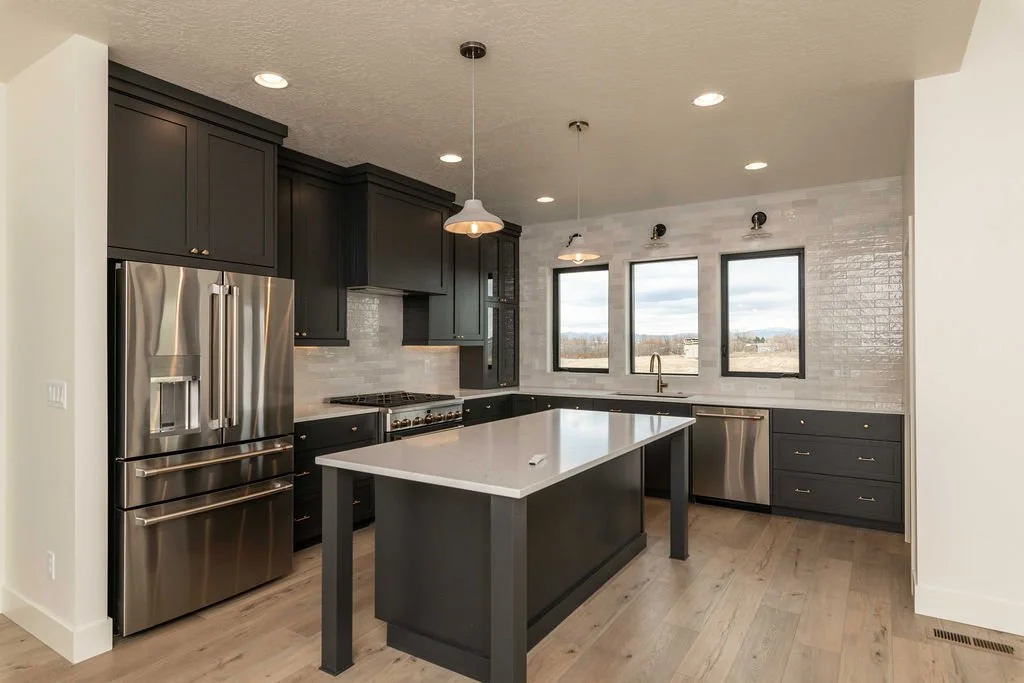 Modern kitchen with dark cabinetry, stainless steel appliances, a central island, three windows above the sink, and white subway tile backsplash.