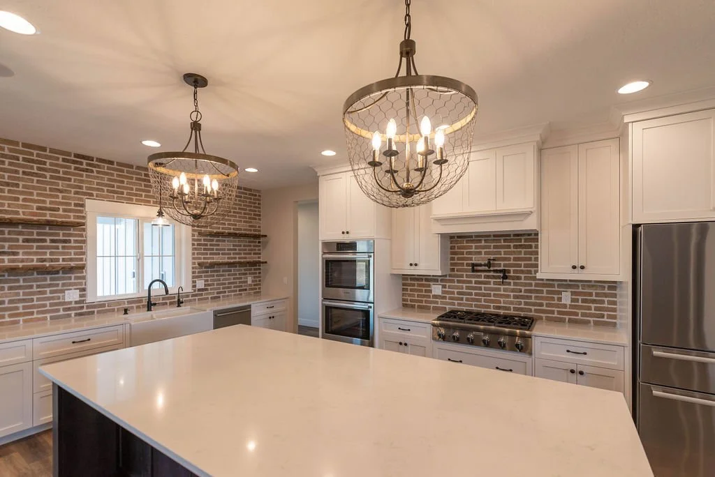 Modern kitchen with white cabinetry, stainless steel appliances, brick backsplash, and a large island with a white countertop. There are two rustic-style chandeliers hanging from the ceiling.