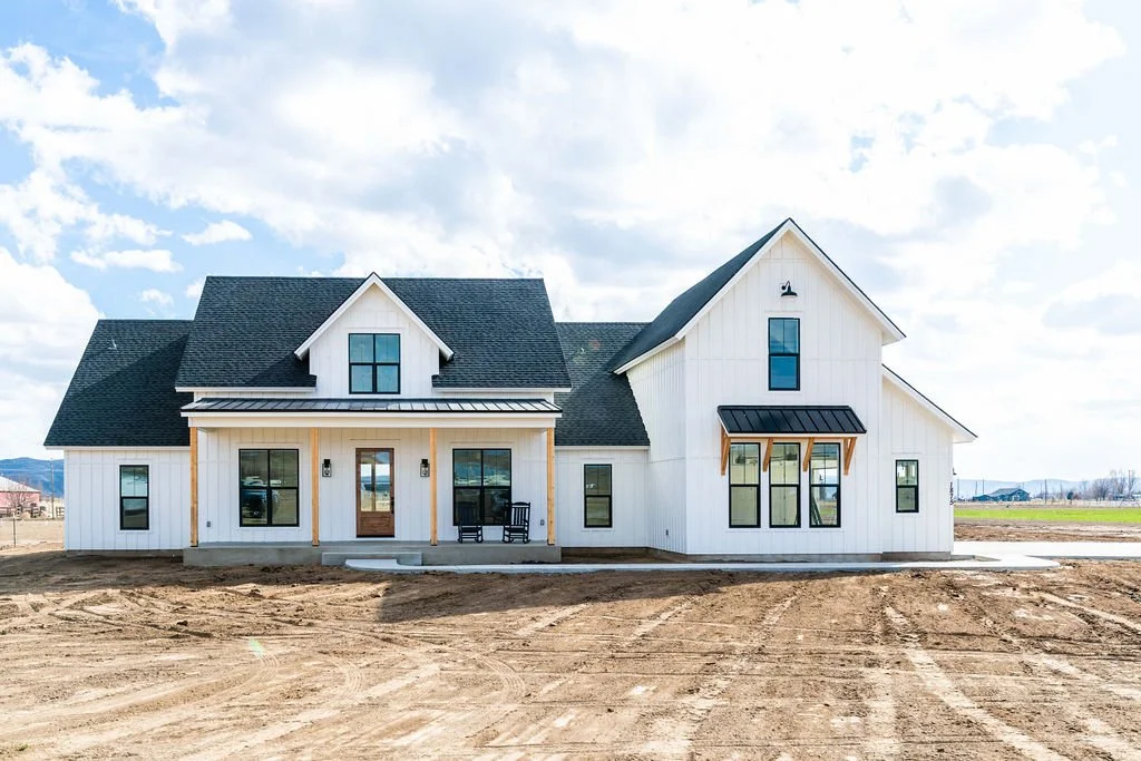 New white farmhouse-style house with black roof, front porch, multiple windows, and a clear sky, under construction on a dirt lot.