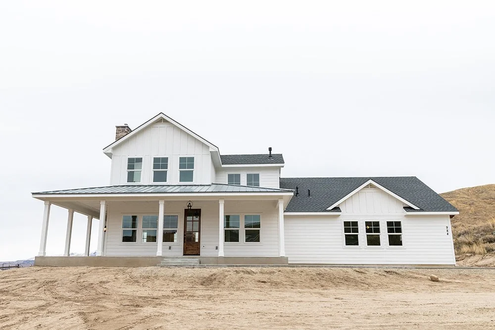 A large white house with a dark gray roof, front porch, multiple windows, and a chimney, surrounded by a dirt yard and an empty landscape.