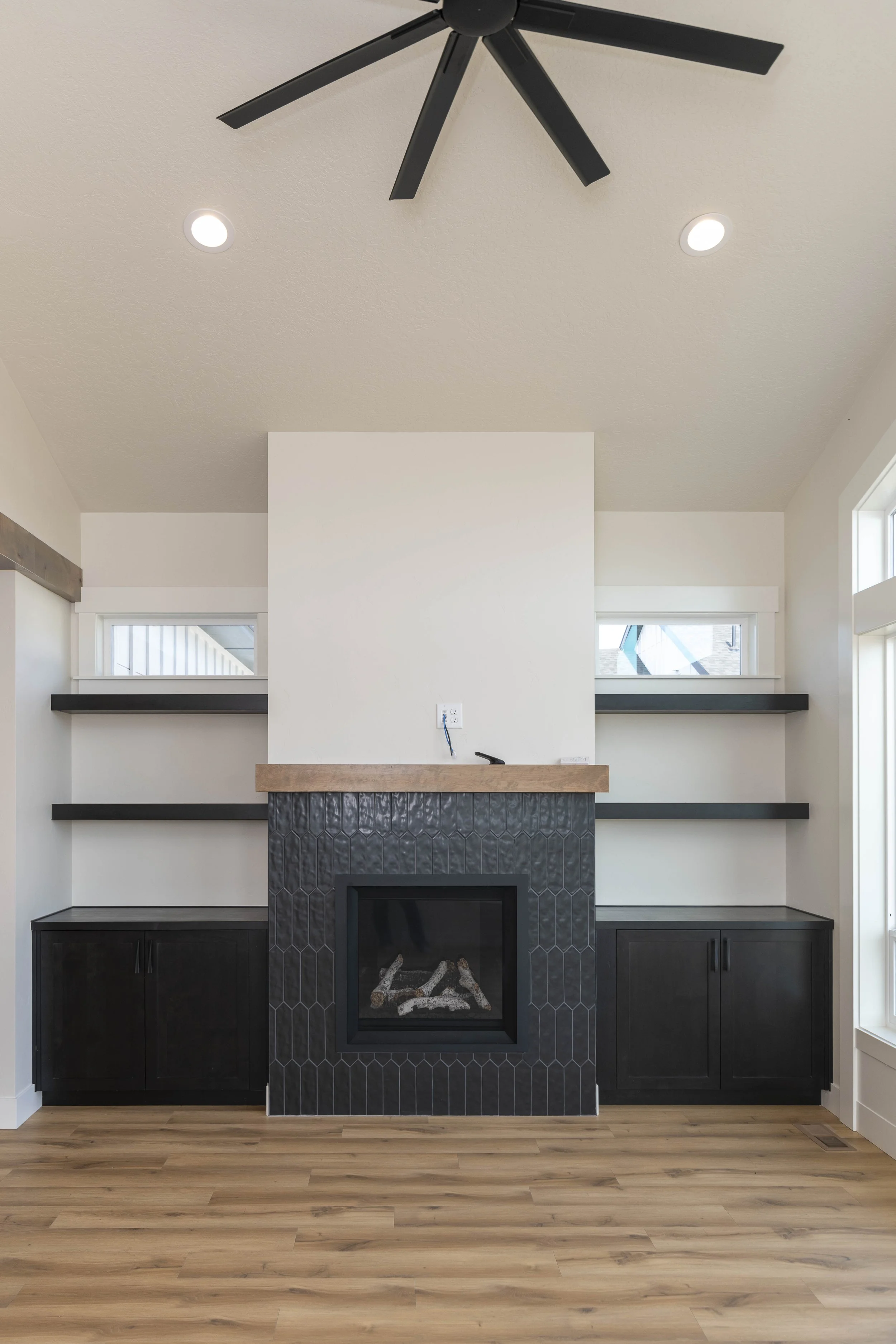 Living room with a black fireplace, black upper and lower cabinets, a half wall with black shelving, and a ceiling fan with black blades, natural light from two high windows, and light wood flooring.