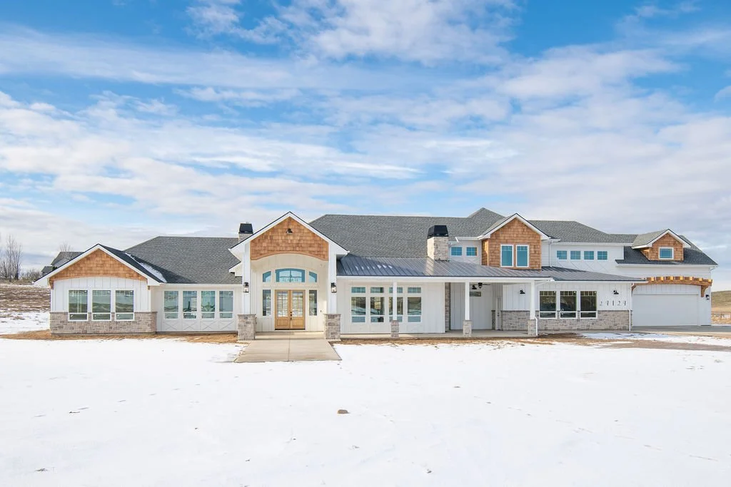 Modern house with a gray roof, white siding, and large windows in a snowy landscape under a partly cloudy sky.