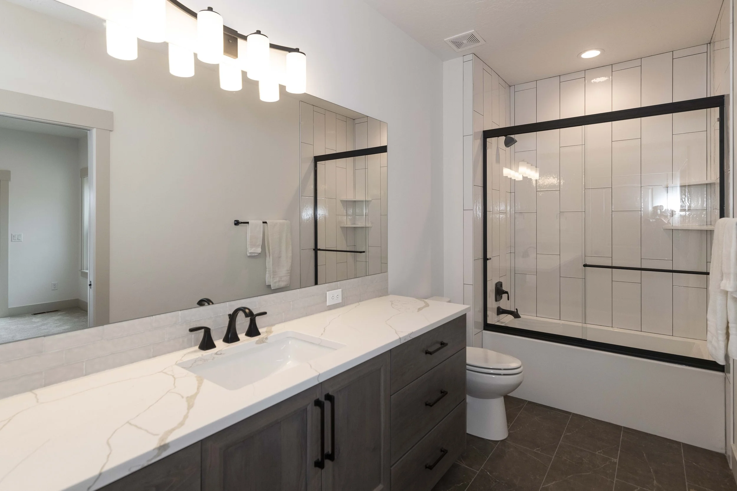 Modern bathroom featuring a large mirror, double sink vanity with dark cabinetry, white marble countertop, towel rack, clear glass shower with black frame, white tiled walls, and a toilet.
