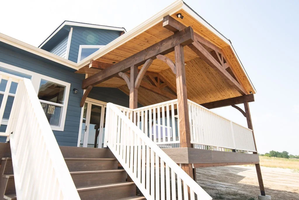 Exterior view of a modern two-story house with a covered porch, blue siding, white railings, and a wooden staircase leading up to it.