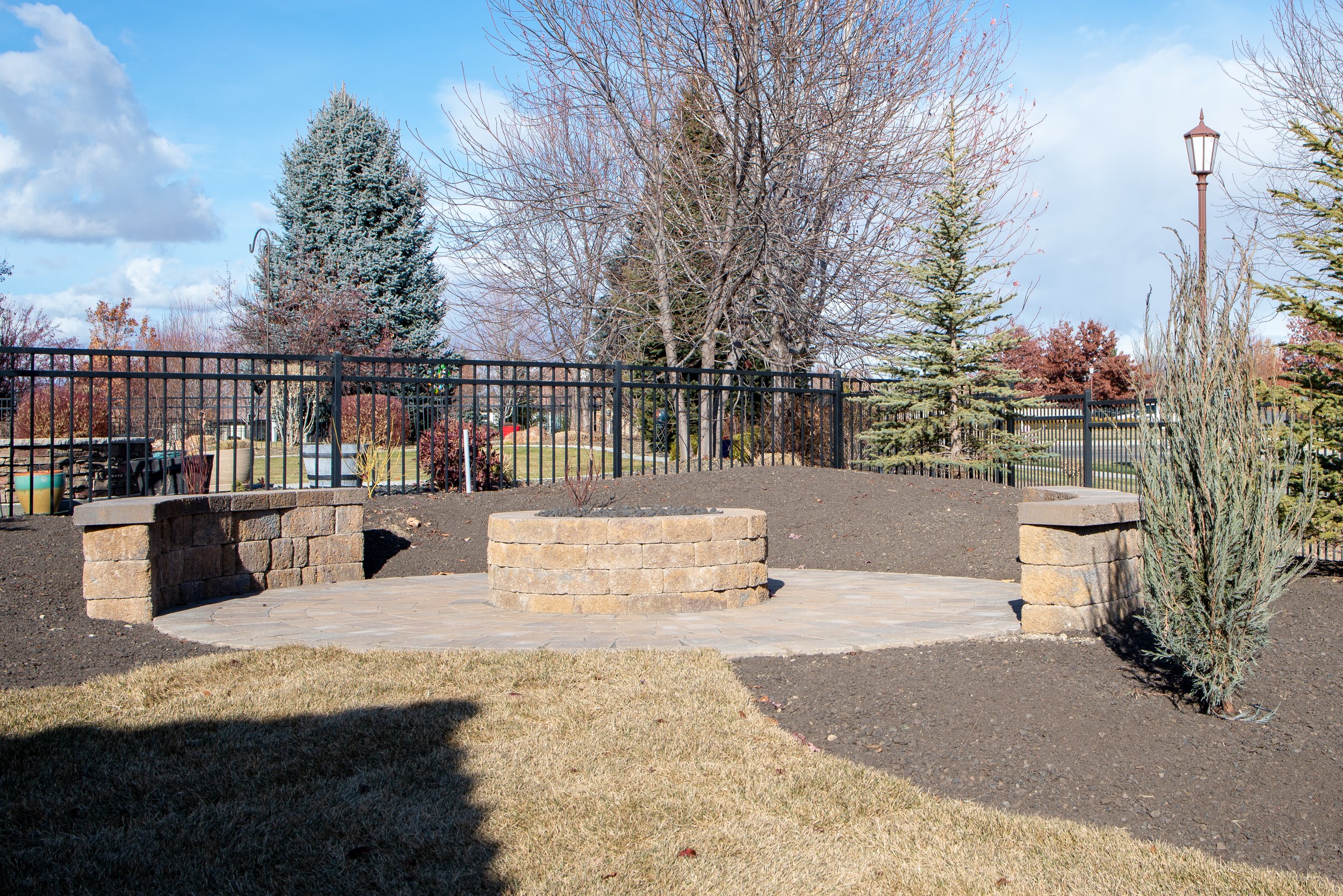 A landscaped outdoor area with a circular stone fire pit, surrounded by a black metal fence. There are trees, including some evergreens and deciduous trees without leaves, and a lamppost. The sky is blue with some clouds.