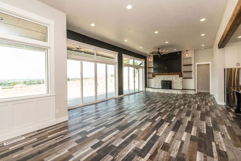 Spacious living room with large windows and sliding glass doors, hardwood floors, a stone fireplace with a black chimney, and built-in shelves on either side of the fireplace.