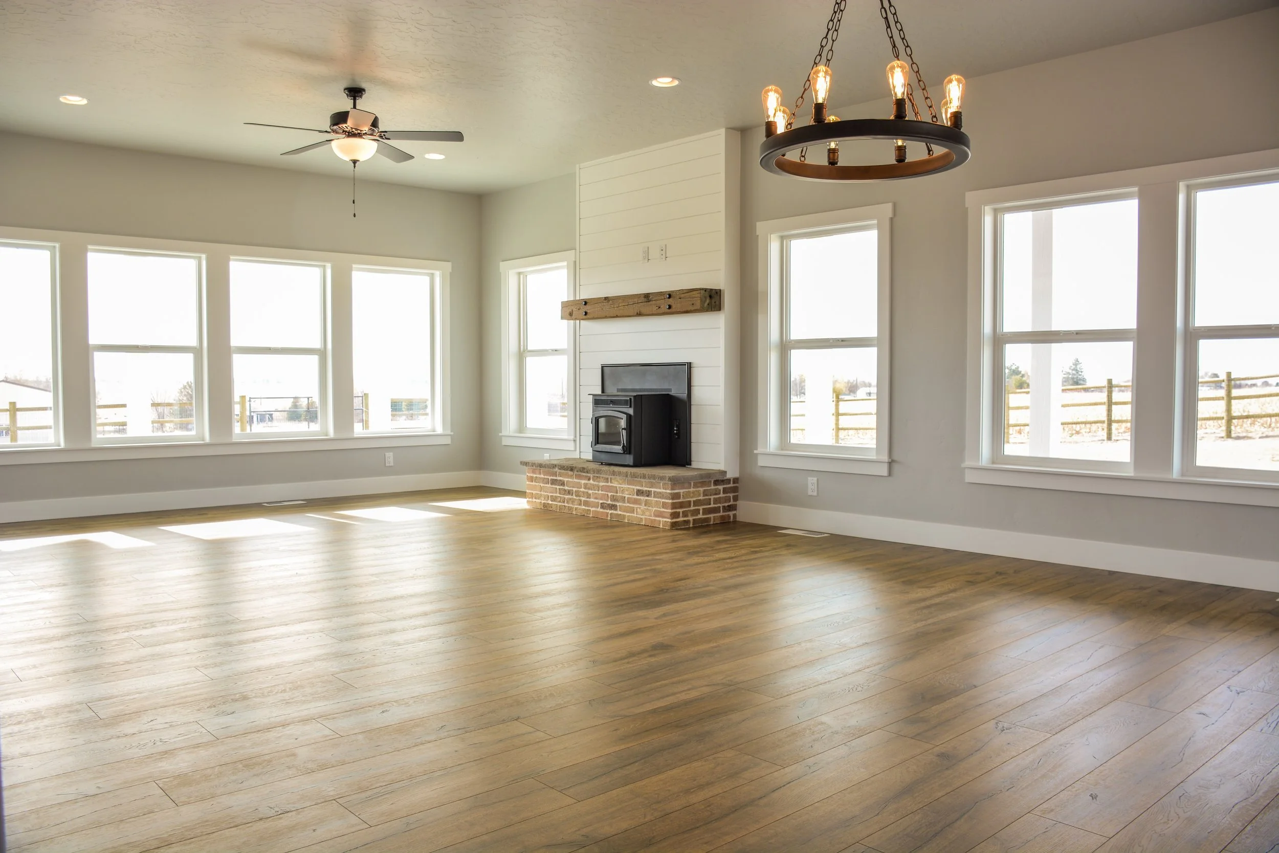 Empty living room with large windows, wooden flooring, a ceiling fan, a chandelier, and a fireplace with a brick base.