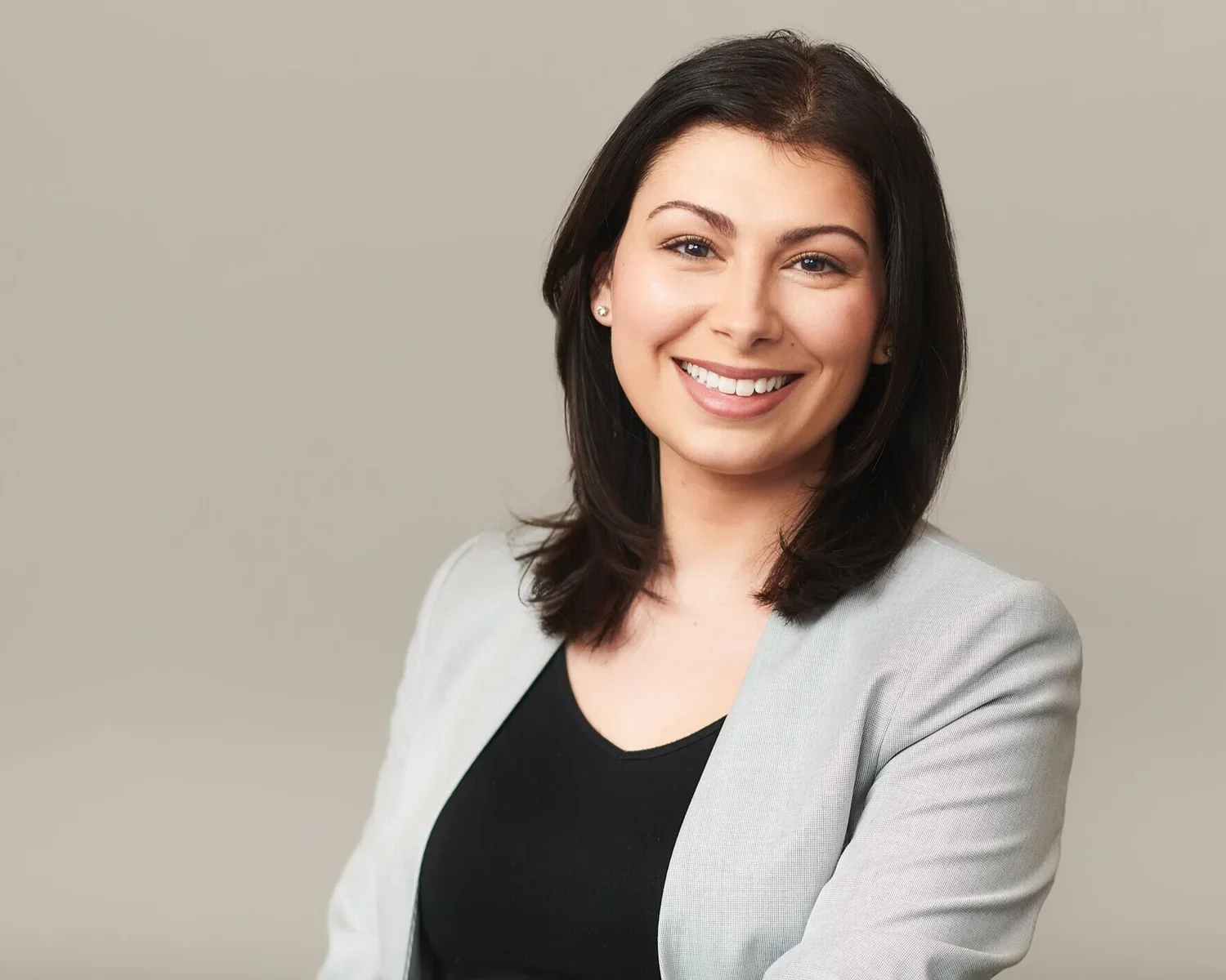 A woman with shoulder-length dark hair smiling, wearing a light gray blazer over a black top, against a plain light gray background.