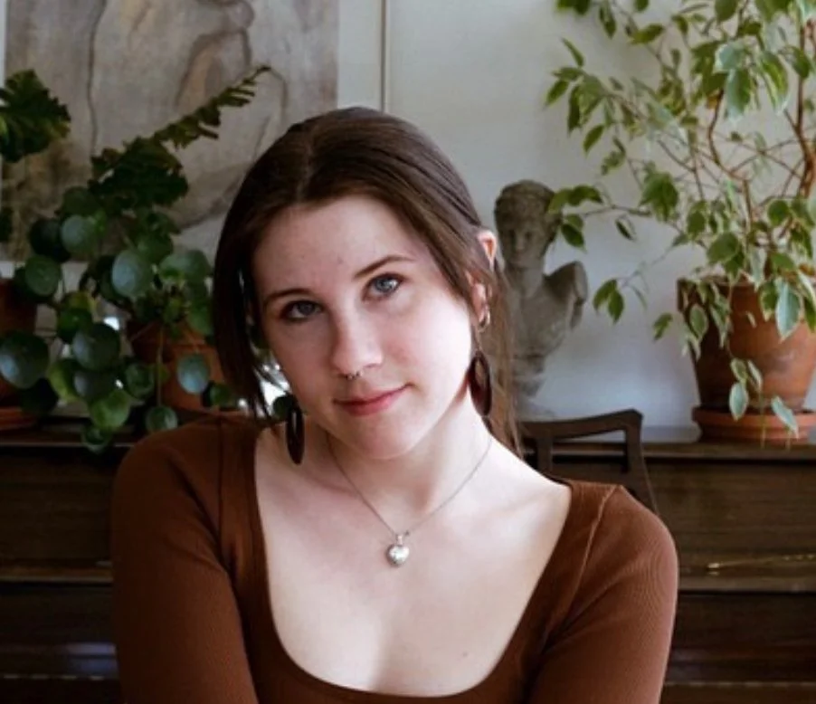 A young woman with brown hair and blue eyes sitting indoors in front of a garden-themed background with potted plants and a stone sculpture.
