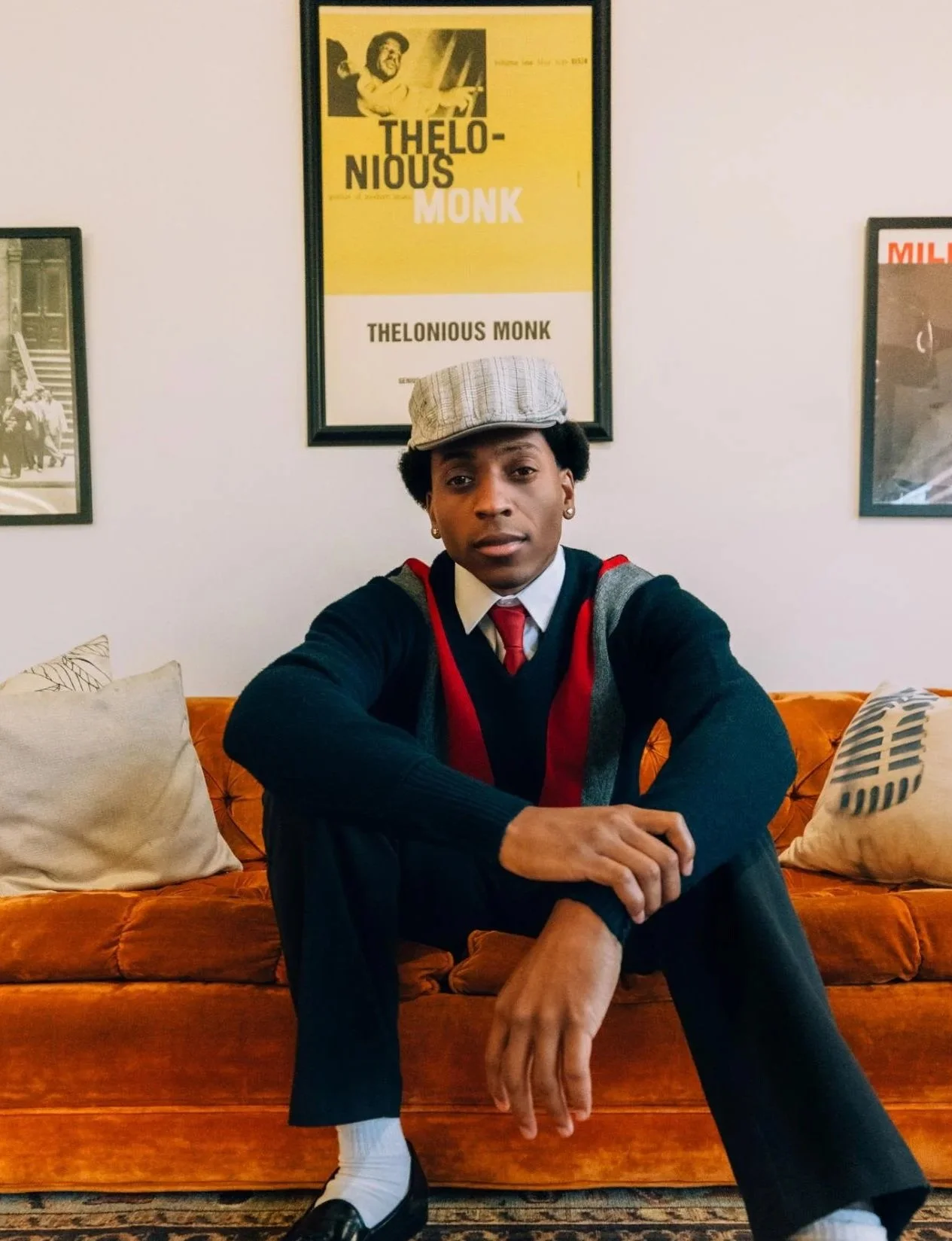 A young man sitting on an orange velvet sofa in a room decorated with framed posters, including one of Thelonious Monk, wearing a gray cap, black sweater with red and gray accents, white shirt with a red tie, black trousers, white socks, and black dress shoes.