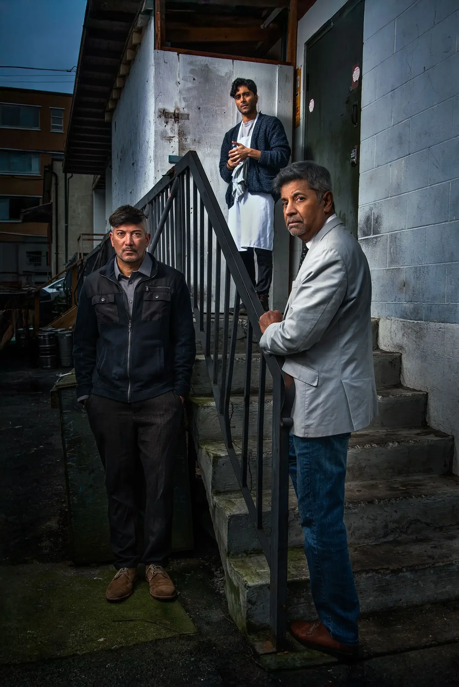 Three men standing on a staircase outside a building at dusk. One man in a white coat and dark hair in the upper part of the staircase, one in a gray jacket with dark hair standing at the bottom next to the staircase, and one in a beige suit with gray hair at the bottom right holding the staircase railing. The building has gray walls, a black door, and a staircase leading upwards. The ground is wet and there is some greenery.