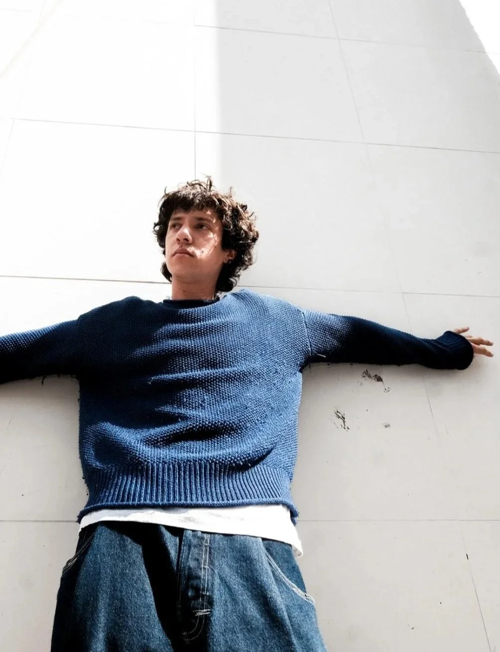 Young man with curly hair wearing a blue sweater and jeans, lying on a tiled white ground with arms outstretched.