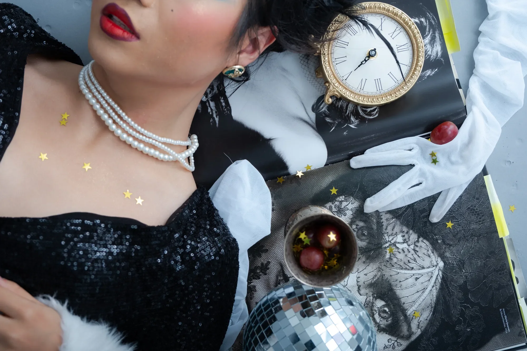 A woman with black hair and red lipstick lying down on a black and white photo print. She wears a pearl necklace and an earring, with gold star stickers on her skin. Nearby are a gold clock, a transparent glove, a glass with red grapes and star-shaped confetti, and a disco ball.