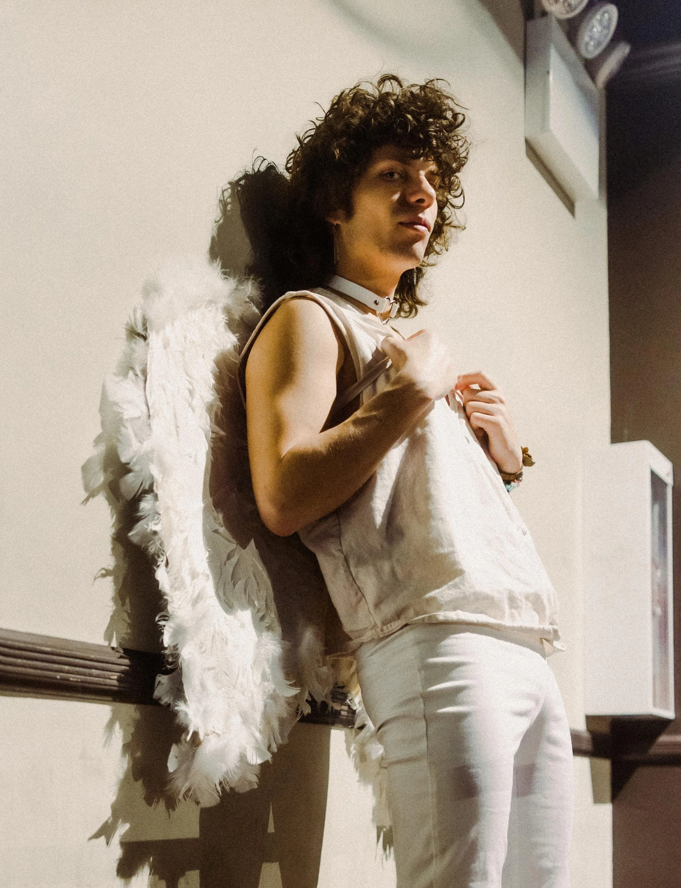 A person with curly hair wearing a sleeveless top, white pants, and angel wings, standing against a plain wall with shadows cast on it.