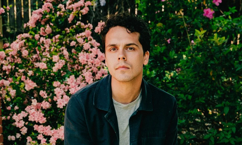 Portrait of a young man with dark curly hair and fair skin, wearing a dark jacket over a light shirt, standing outdoors in front of pink flowering bushes and green foliage.