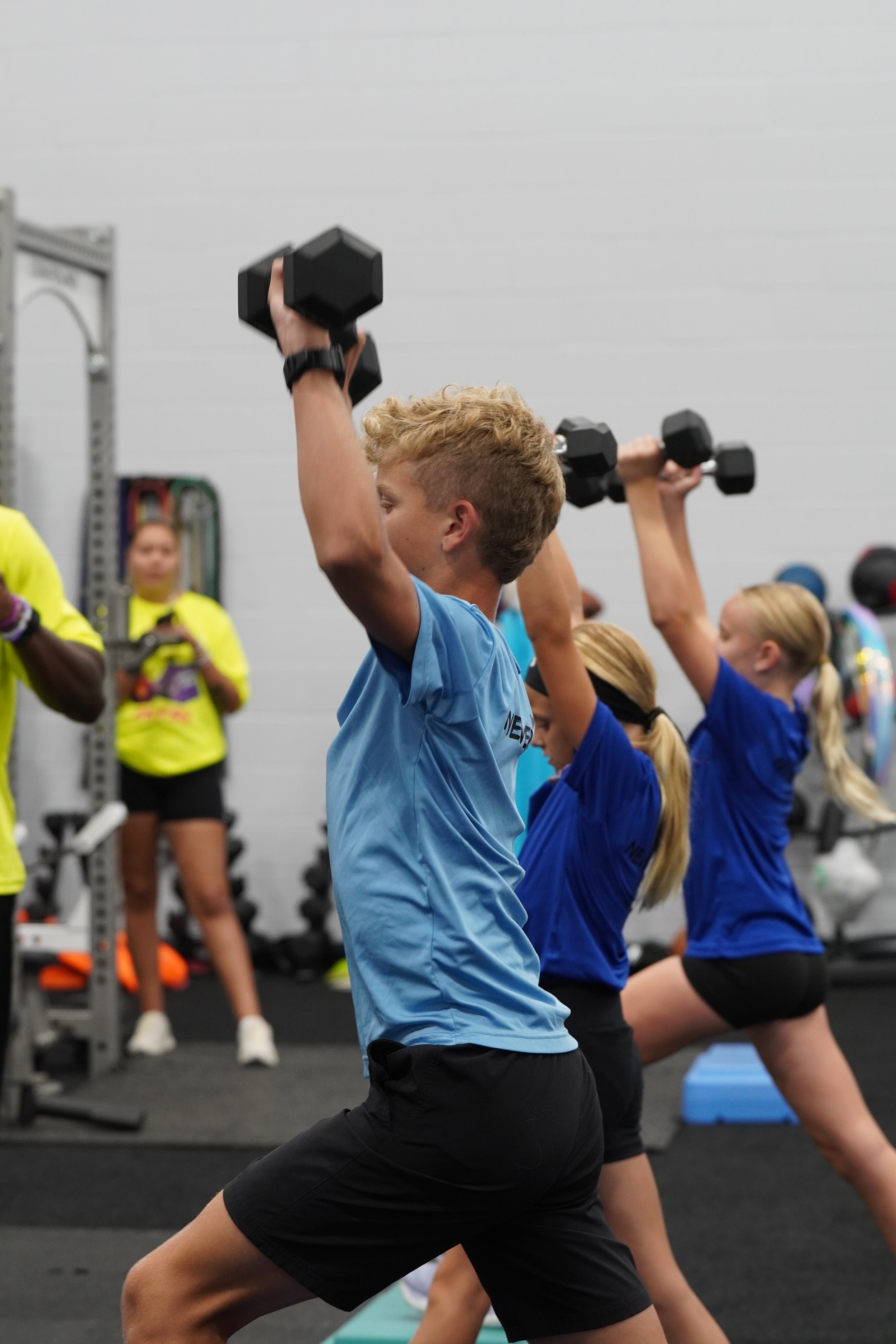 A group of people, including children and adults, are exercising indoors. They are performing a yoga pose with one leg bent and one arm extended, holding a dumbbell overhead. The setting appears to be a gym or fitness class with equipment and other participants in the background.