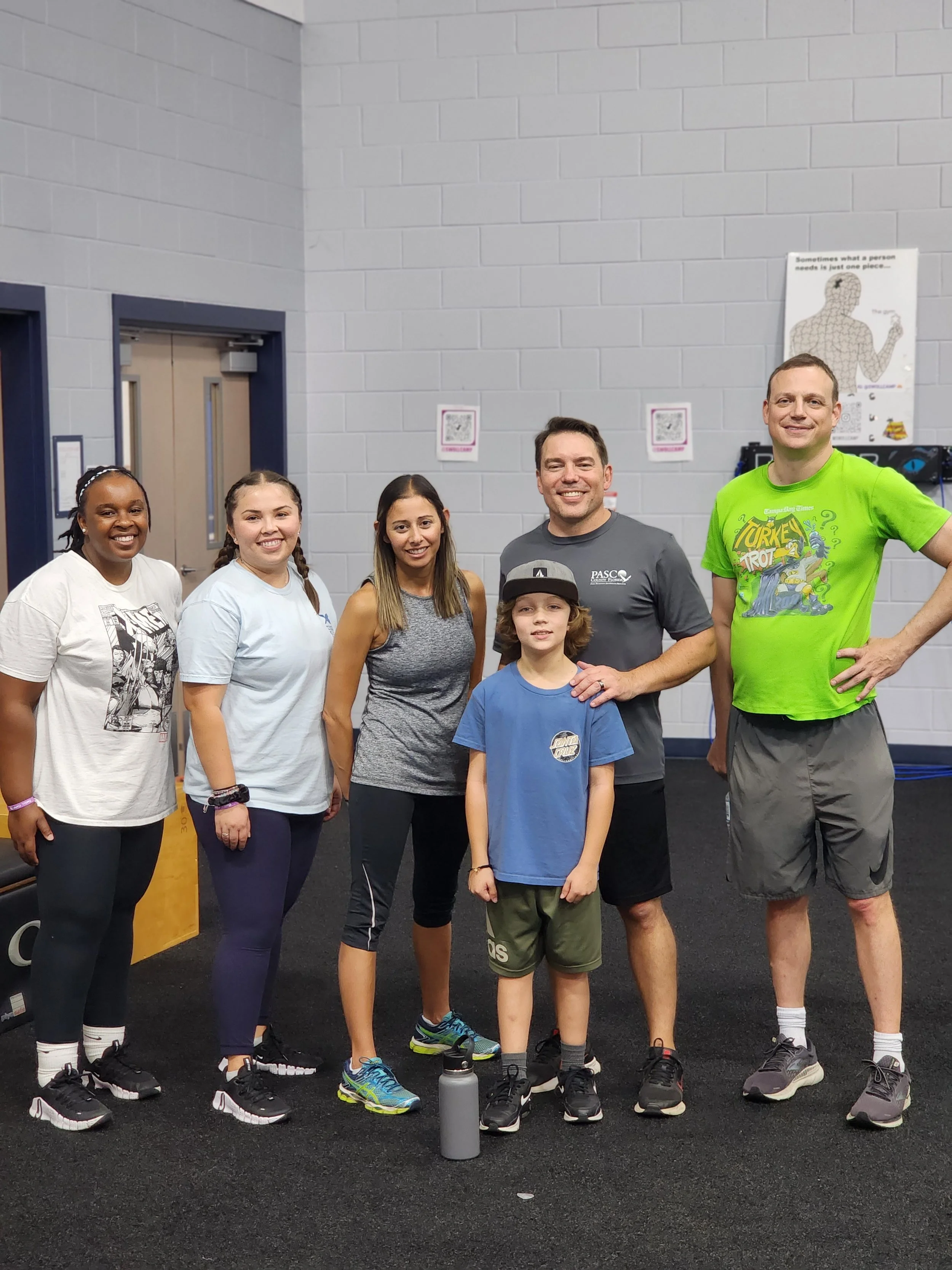 A group of seven people, including two children, standing together inside a gym or fitness center, smiling at the camera, casually dressed in athletic clothing, with water bottles on the floor in front of them.