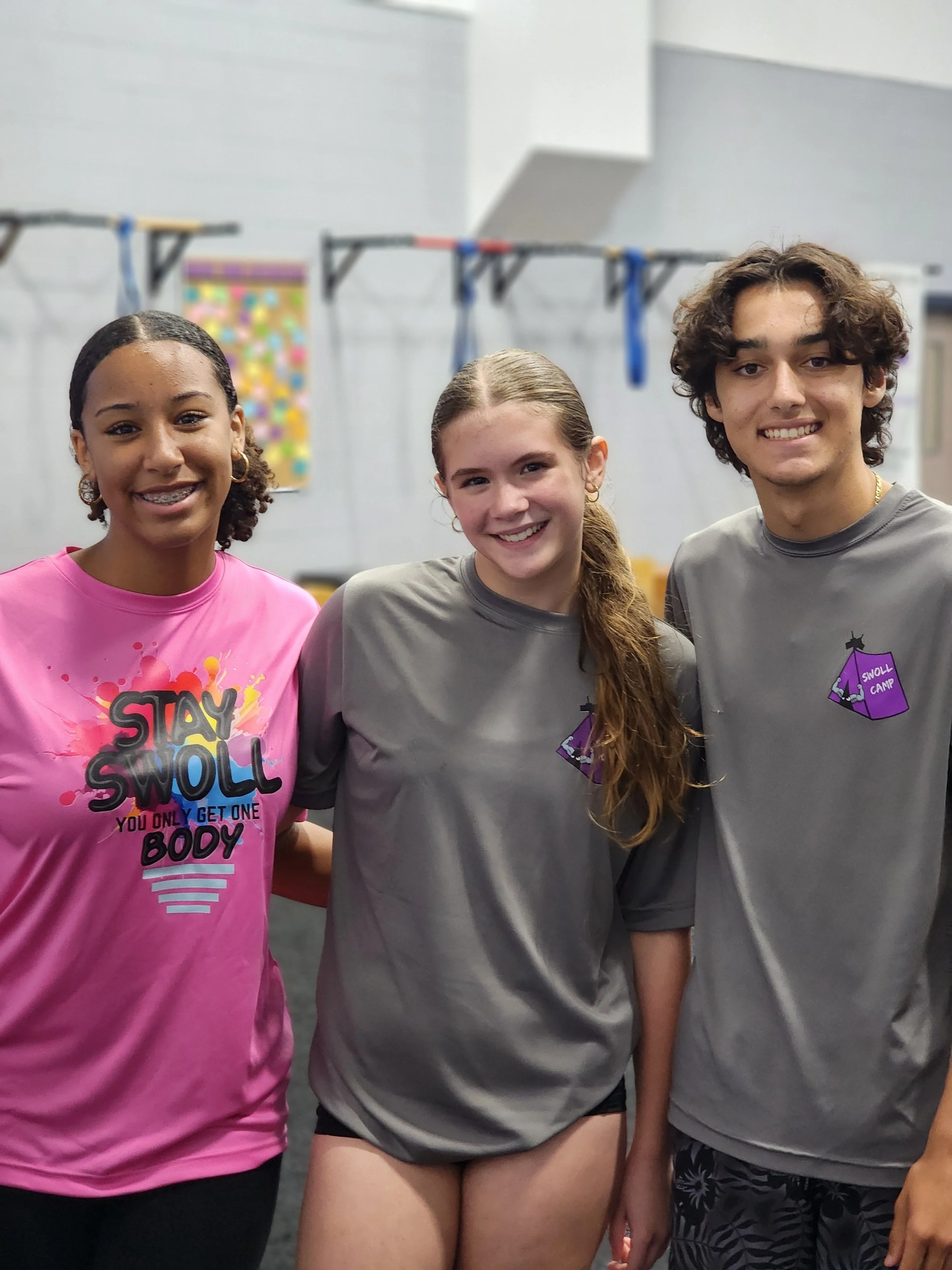 Three children standing side by side indoors. The girl on the left is wearing a pink shirt with colorful text, the girl in the middle is in a gray shirt, and the boy on the right is in a gray shirt with patterns. They are smiling at the camera.
