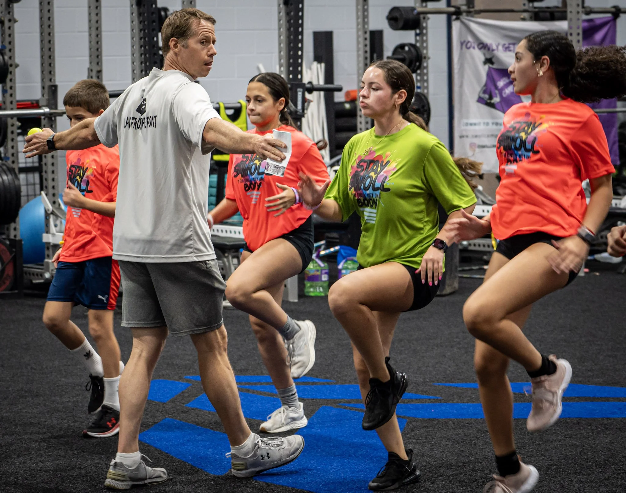 A group of people participating in a fitness class or workout in a gym, doing synchronized exercises on a rubber floor, with a man leading and others following, wearing colorful workout shirts.