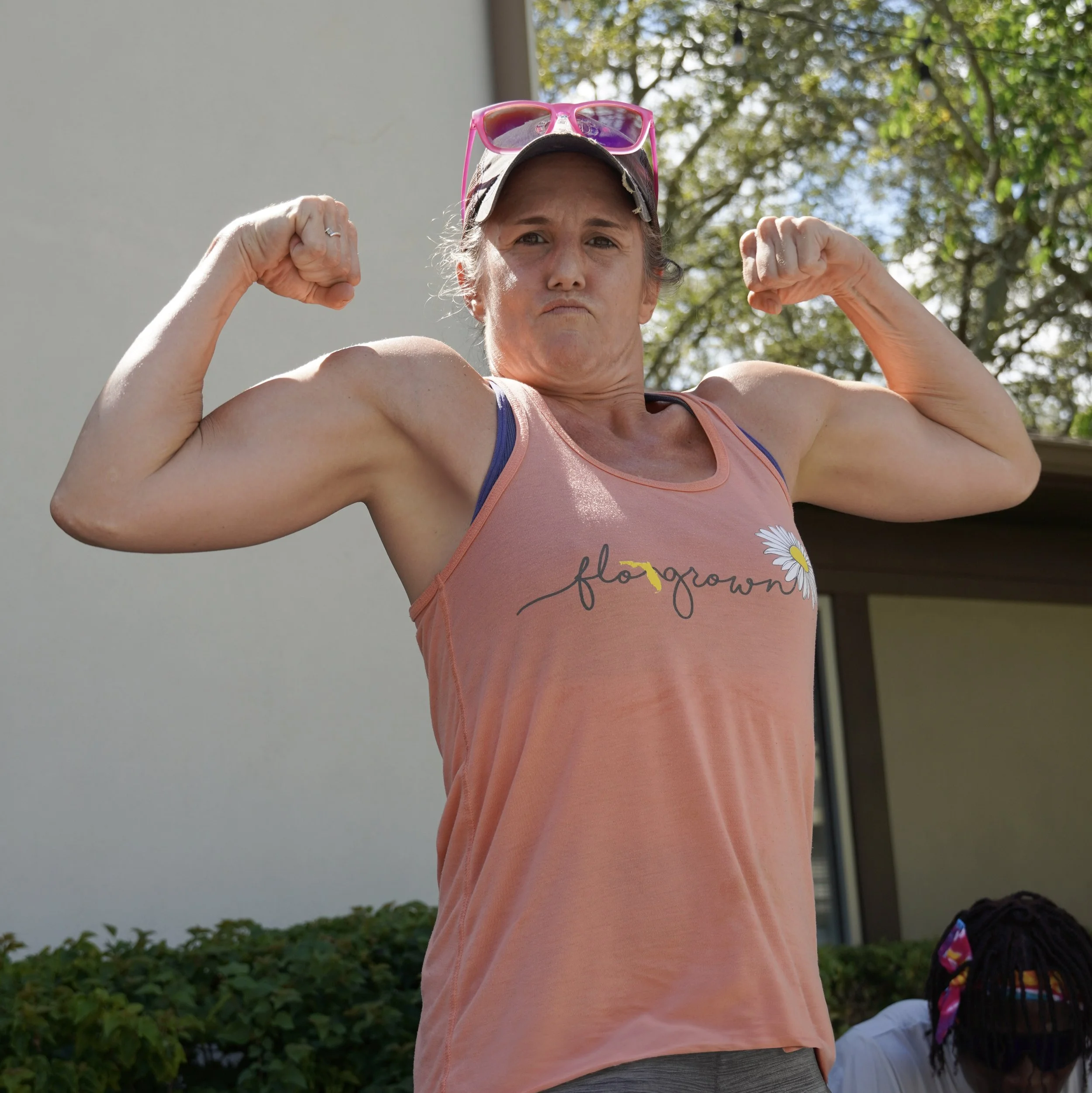 A woman with sunglasses on her head flexing her biceps outdoors, wearing a pink tank top with the word 'flower' and daisies printed on it.
