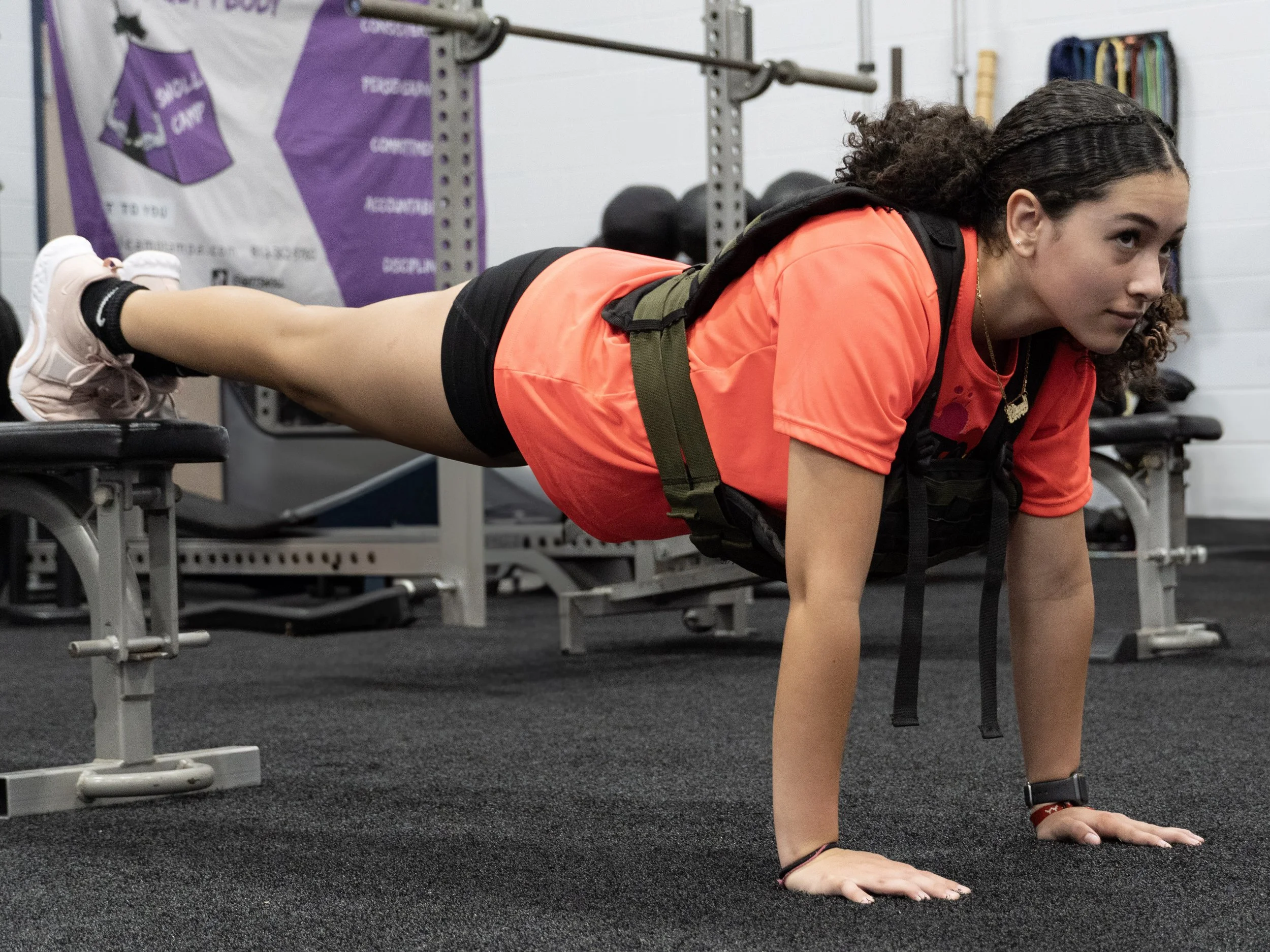 A young woman with curly hair doing a plank exercise on a black gym floor, in a gym setting with weights and fitness equipment in the background.