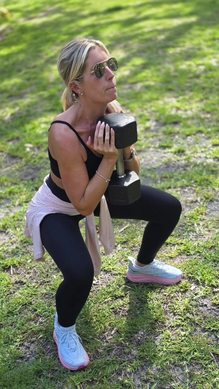 A woman performing a squat outdoors while holding a dumbbell in each hand, wearing sunglasses, a black tank top, black leggings, white sneakers with pink soles, and a light-colored jacket tied around her waist.
