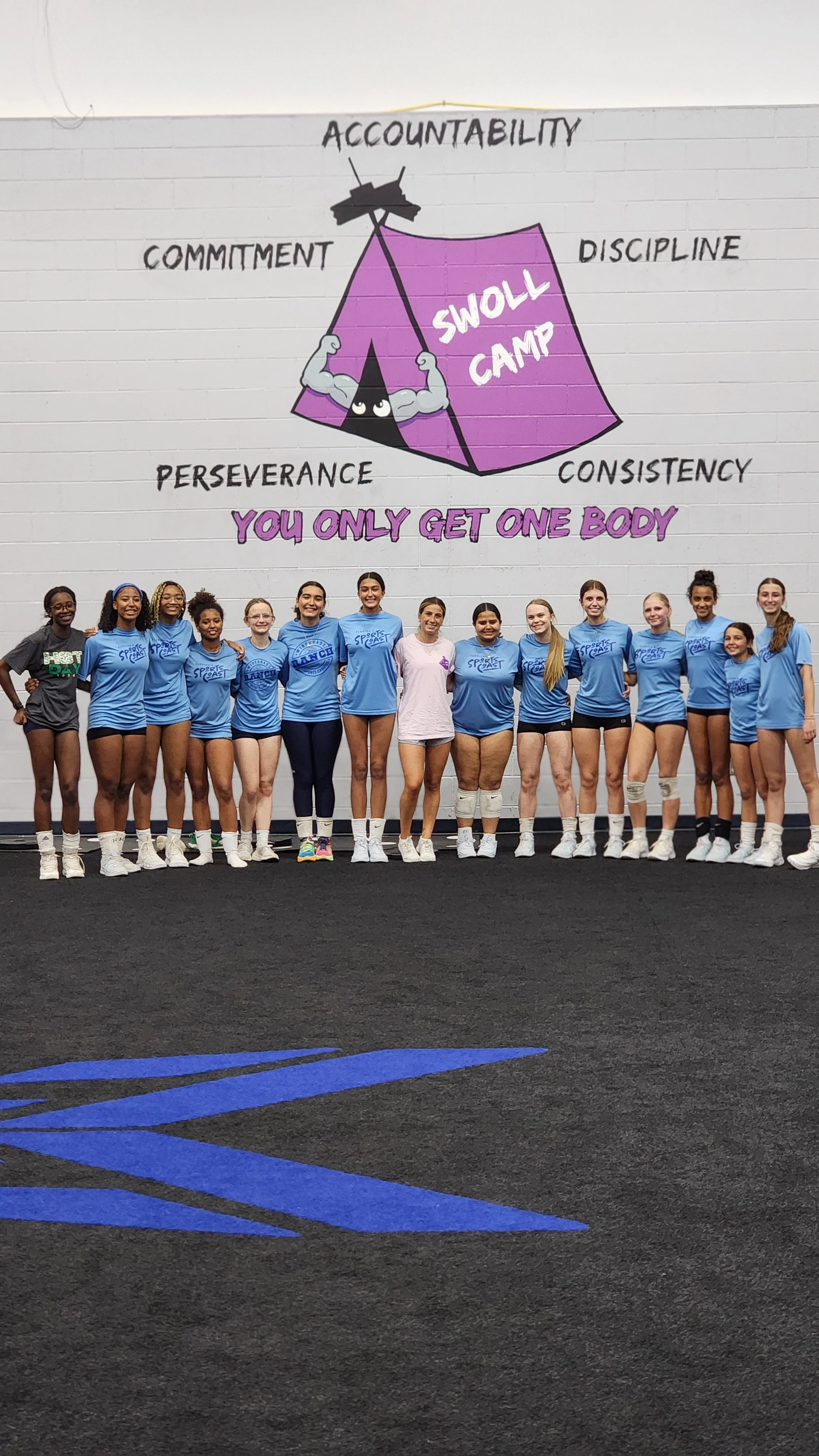 A group of young female cheerleaders standing in front of a large motivational wall poster. The poster features a purple tent with a muscular character and words like 'accountability,' 'commitment,' 'perseverance,' 'discipline,' and 'consistency.' The phrase 'You only get one body' is written at the bottom of the poster.