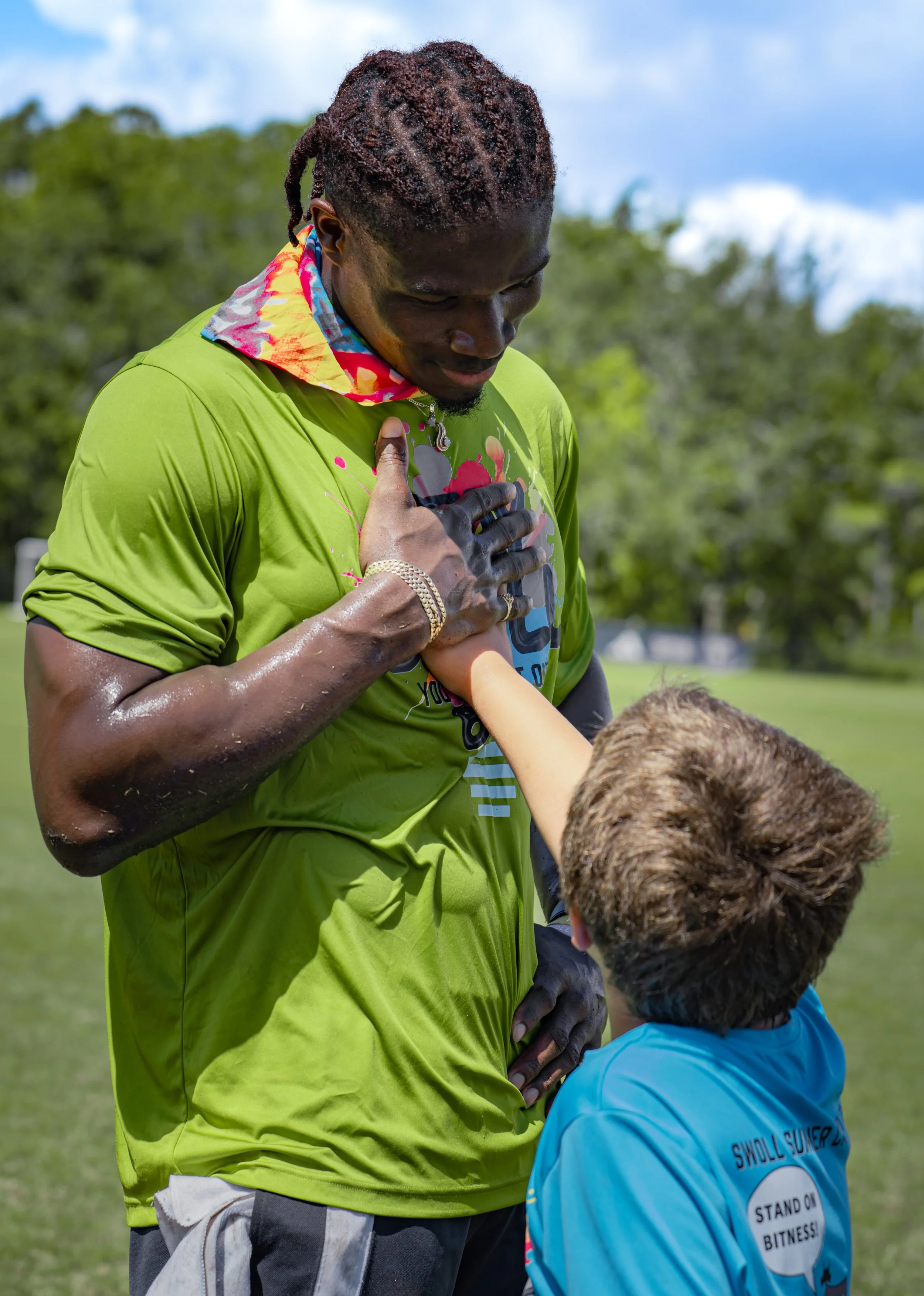 A man with dreadlocks, wearing a bright green shirt and colorful neck gaiter, places his hand over his heart and looks down at a young boy, who is wearing a blue shirt and standing in front of him outdoors with trees and a cloudy sky in the background.