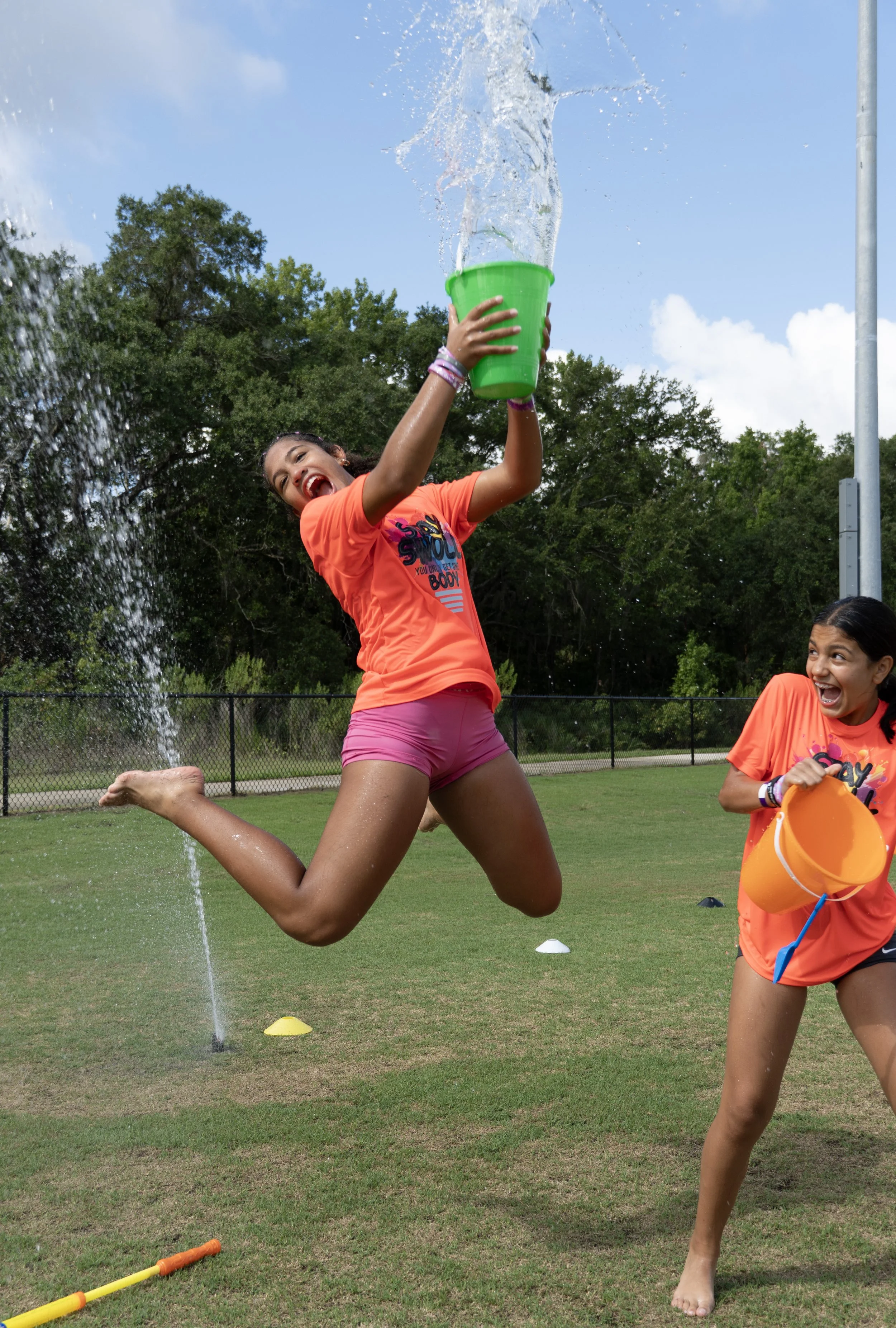 Two young girls playing outside on a grassy field during daytime. One girl with dark hair, wearing a bright orange T-shirt, pink shorts, and a bracelet, is jumping near a sprinkler with water splashing around. She is holding a green bucket, and another girl, also wearing an orange T-shirt, is smiling and holding an orange bucket. The background features trees and a blue sky with some clouds.