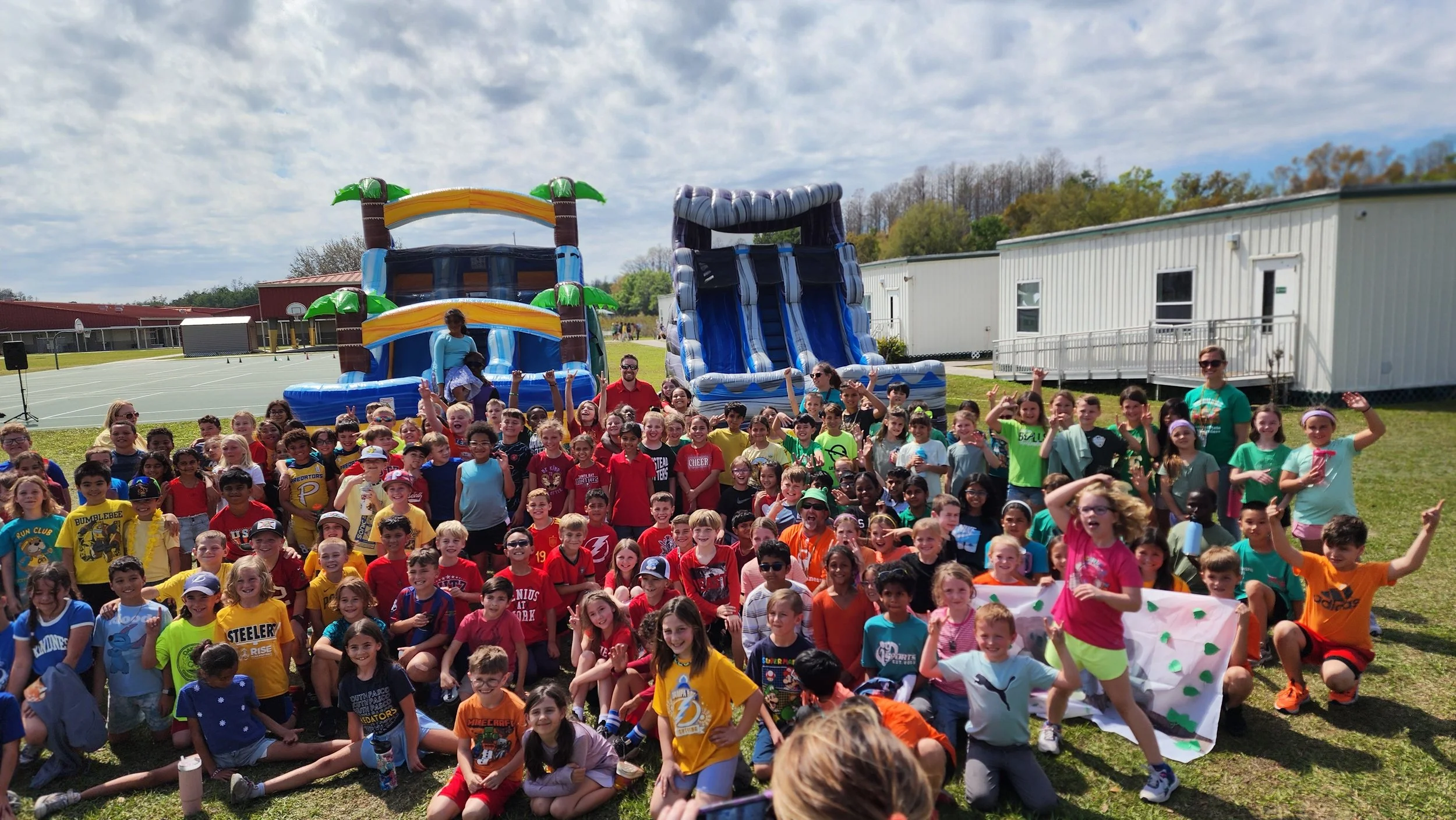 Large group of children gathered outdoors in front of inflatable slides, posing for a group photo under a partly cloudy sky.