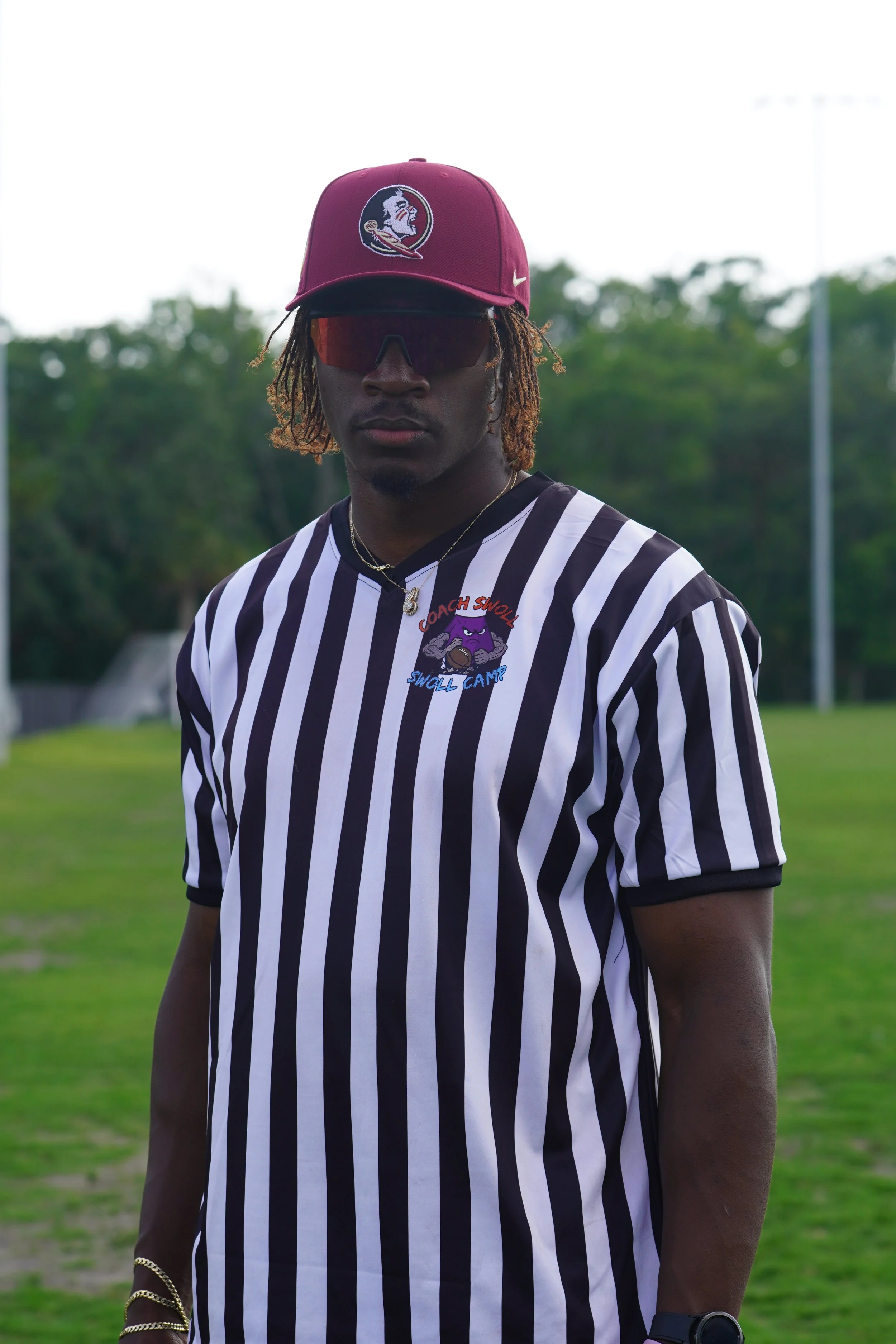 A young man wearing a maroon cap with a logo, reflective sunglasses, a black and white striped sports jersey, and gold jewelry standing outdoors on a grassy field with trees in the background.
